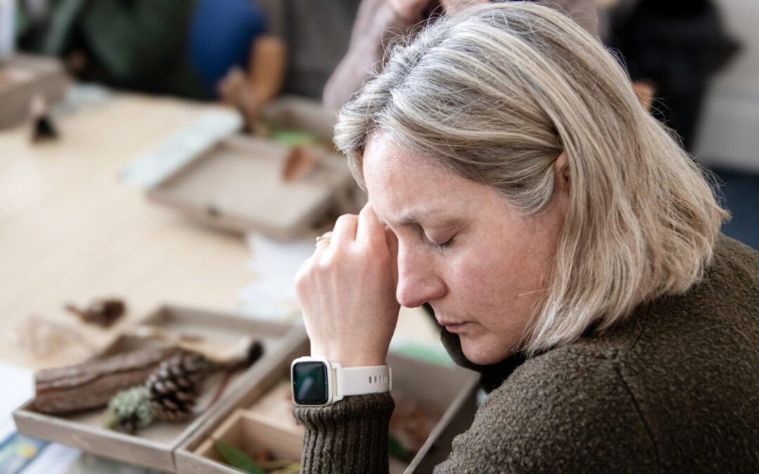 Nature indoors. A lady holding some leaves to her ear listening to them make a crunching sound. Has a wooden box on the table with forest items in it.